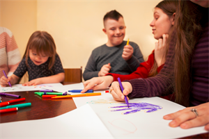 Children and adults sitting at a table colouring hand-prints on paper with pens
